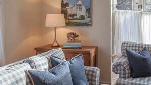 A cozy living room with a checkered sofa, a lamp, a painting of a house, and a window with curtains. - Summerland Stagecoach Cottage - A Historic Treasure in Summerland (Summerland)