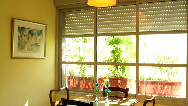 Un coin repas avec une table et des chaises, une fenêtre avec des stores et un tableau encadré au mur.