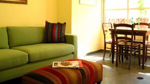 Un salon avec un canapé vert, un coussin rayé aux couleurs vives, un plaid à motifs, une table à manger avec des chaises et une fenêtre avec des stores.