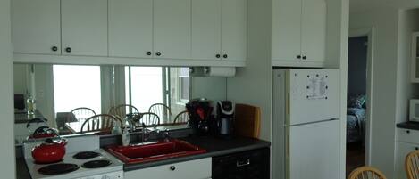 Cocina compacta con armarios blancos, un lavamanos rojo, estufa y lavavajillas negro. Hay sillas de madera alrededor de una pequeña mesa de comedor.