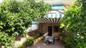 Zona de pati coberta amb taula i cadires, envoltada de plantes en testos i una paret de pedra.