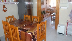 Zona de menjador amb taula i cadires de fusta, un frigorífic i una cuina visible a través d'una obertura.