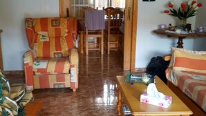 Un saló amb una butaca estampada, una taula de centre de fusta plena de llibres, un sofà, un rellotge a la paret i una porta que dona a una altra estança.