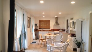 Une cuisine moderne avec des meubles blancs, un coin repas doté de chaises en osier et une grande fenêtre agrémentée de rideaux.