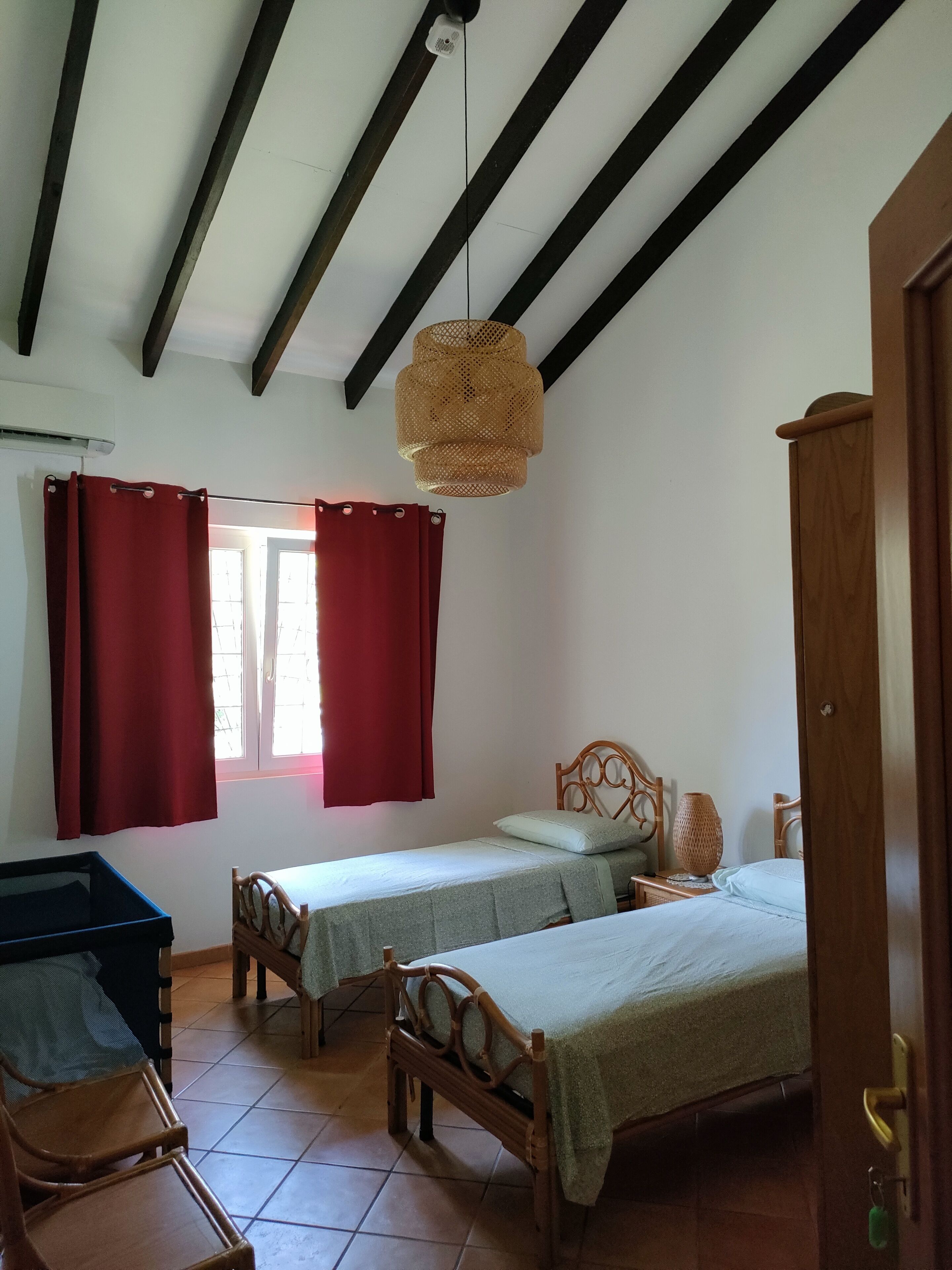 Ein Schlafzimmer mit zwei Einzelbetten, einem Holz-Kopfteil, einer hängenden Gewebedecke und einem Fenster mit roten Vorhängen.