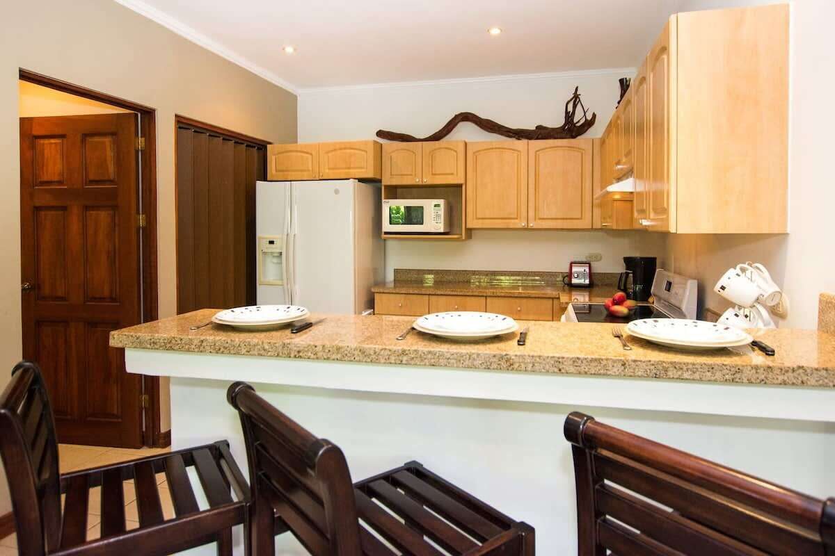 A kitchen with wooden cabinets, a white refrigerator, a microwave, and a granite countertop. There are two wooden chairs and a small island with a sink.