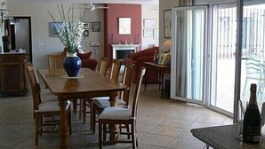 Comedor con mesa y sillas de madera, un florero con plantas, un ventilador de techo y vistas a través de un ventanal.
