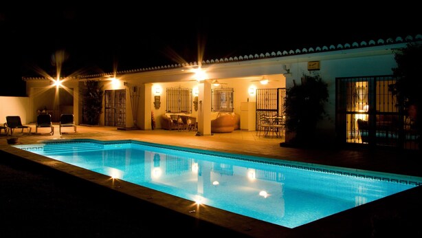 Un área de piscina nocturna con piscina cristalina, sillones de descanso y una zona de estar cubierta con mesas.