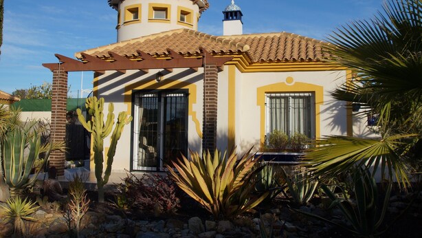 Un edificio de estilo tradicional con una torre circular, una pérgola y una variedad de plantas desérticas.