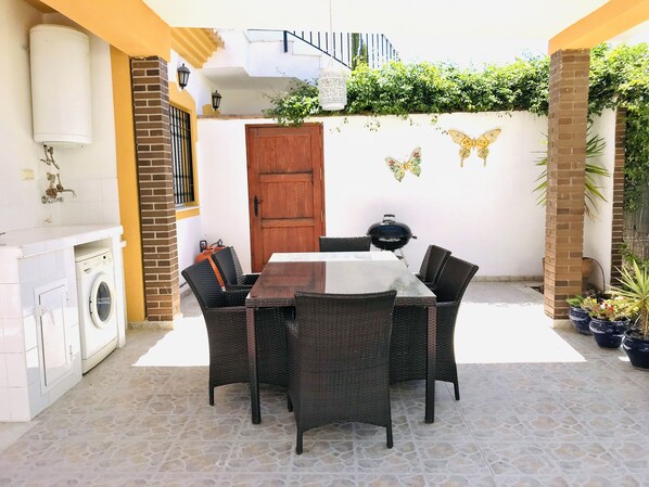 Un patio con mesa y sillas, lavadora, puerta de madera y pared de ladrillo decorada con motivos de mariposas.