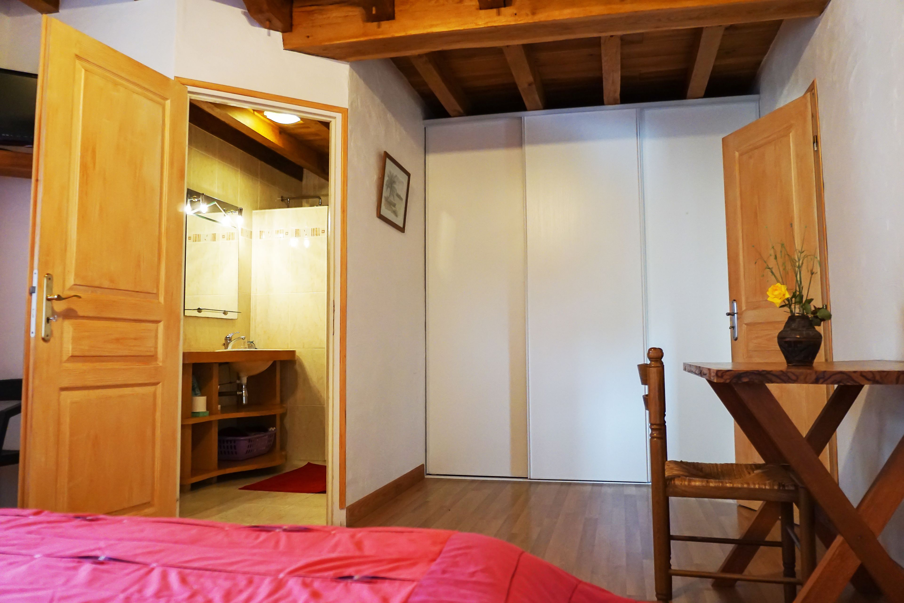 A bedroom with a wooden bed, a wooden chair, a small table with a vase, a wardrobe, and a bathroom with a shower.