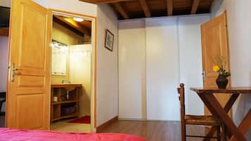 A bedroom with a wooden bed, a wooden chair, a small table with a vase, a wardrobe, and a bathroom with a shower.