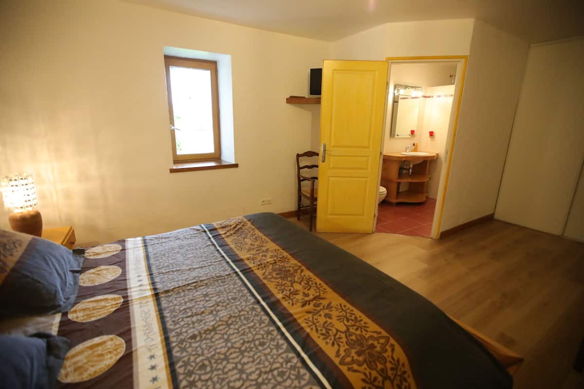 A bedroom with a bed, a window, a door leading to a bathroom, and a bedside table with a lamp.