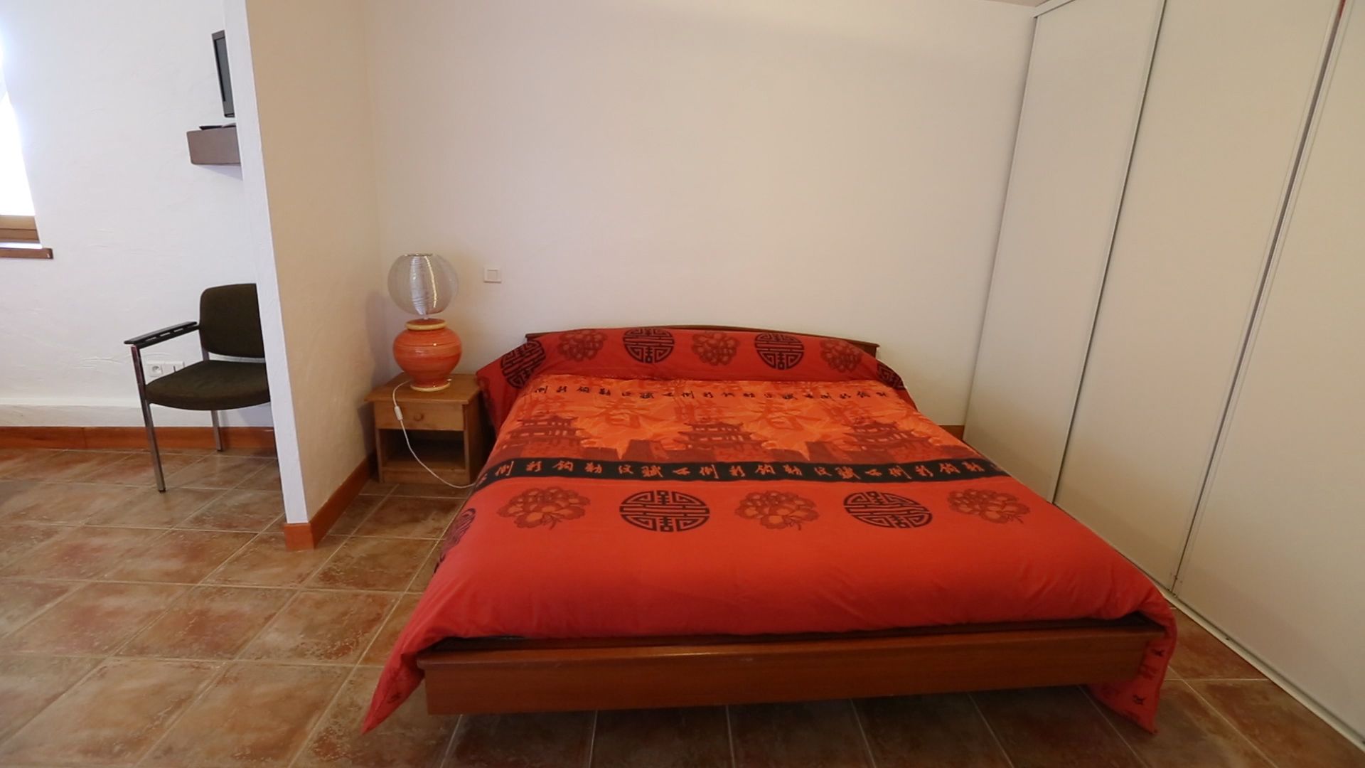 Un dormitorio con cama, una silla, una mesita de noche y un estante en la pared.