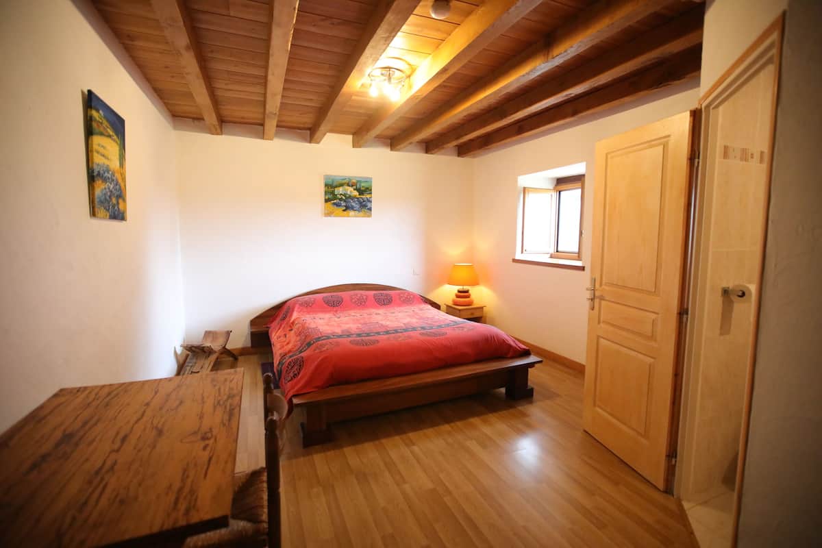 A bedroom with a wooden bed, a nightstand with a lamp, a wooden door, and a window.