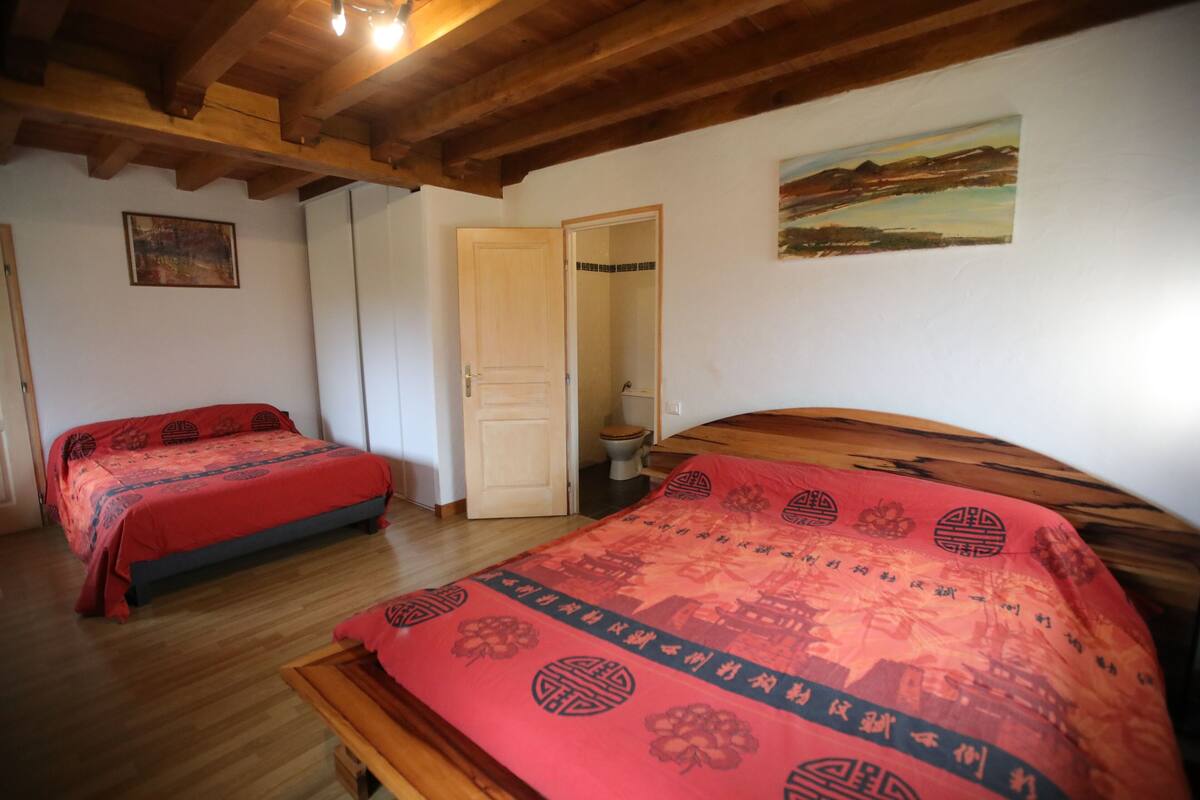 A room with two beds, wooden beams on the ceiling, a painting on the wall, and a door leading to a bathroom.