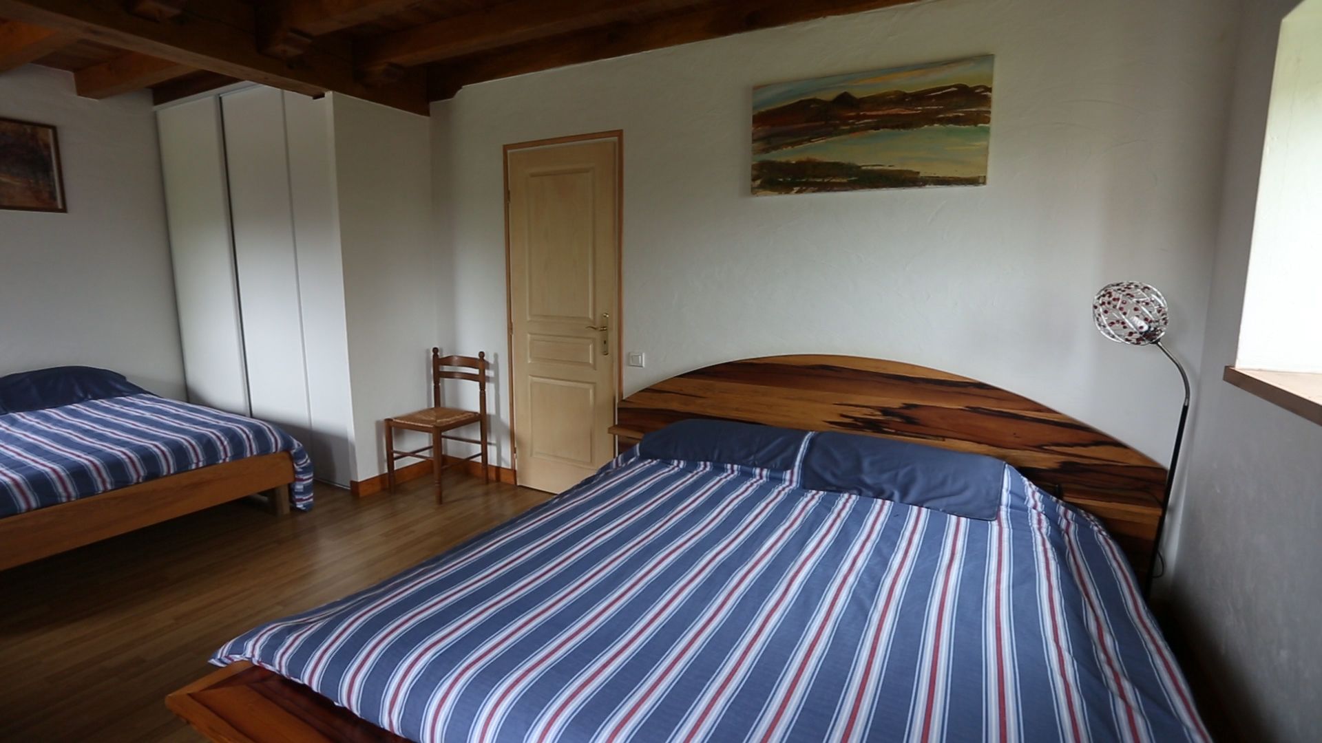 A room with two single beds, a wooden headboard, a chair, a door, and a painting on the wall.