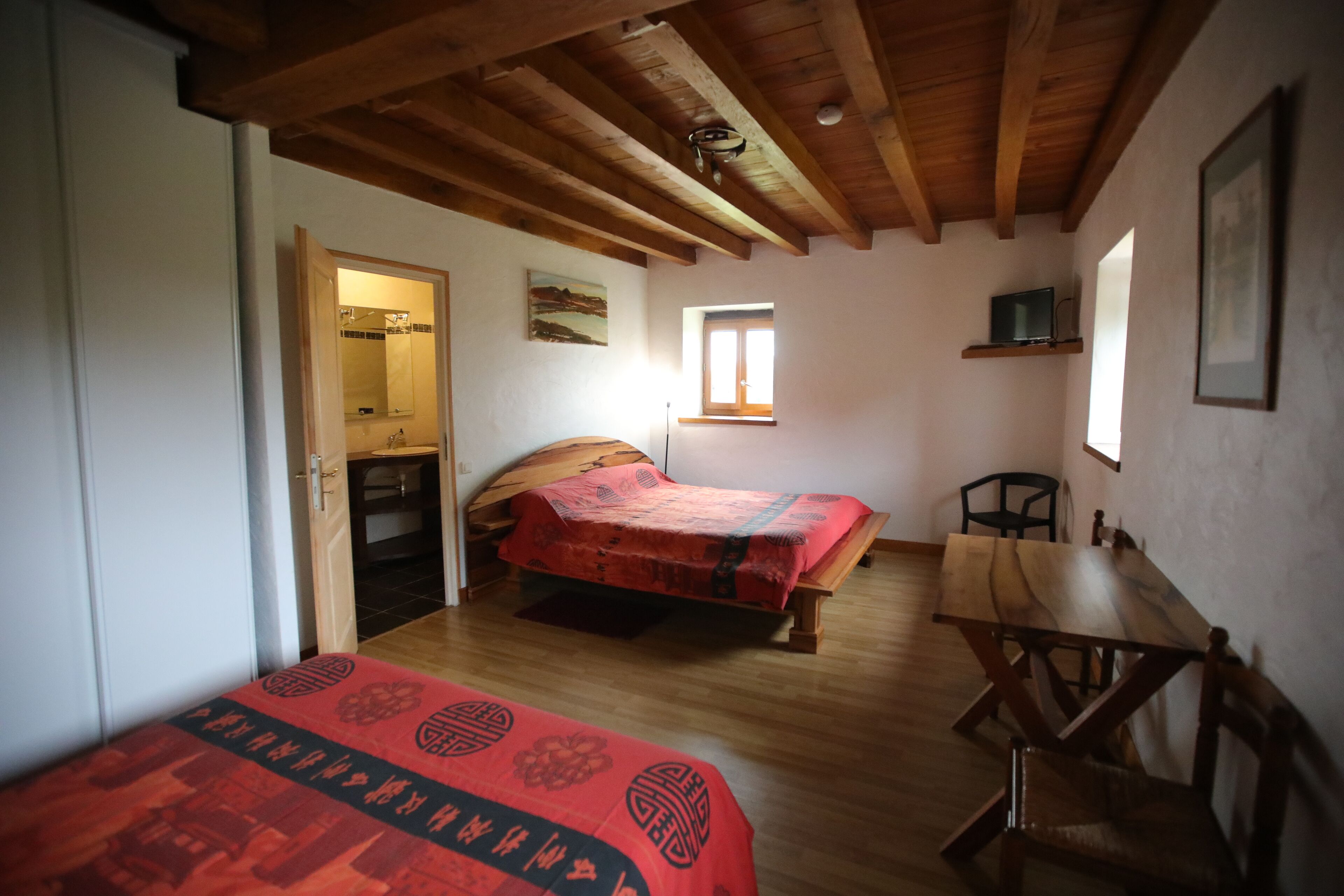 A bedroom with two beds, a wooden table, and a chair. There is a window providing natural light and a door leading to a bathroom.