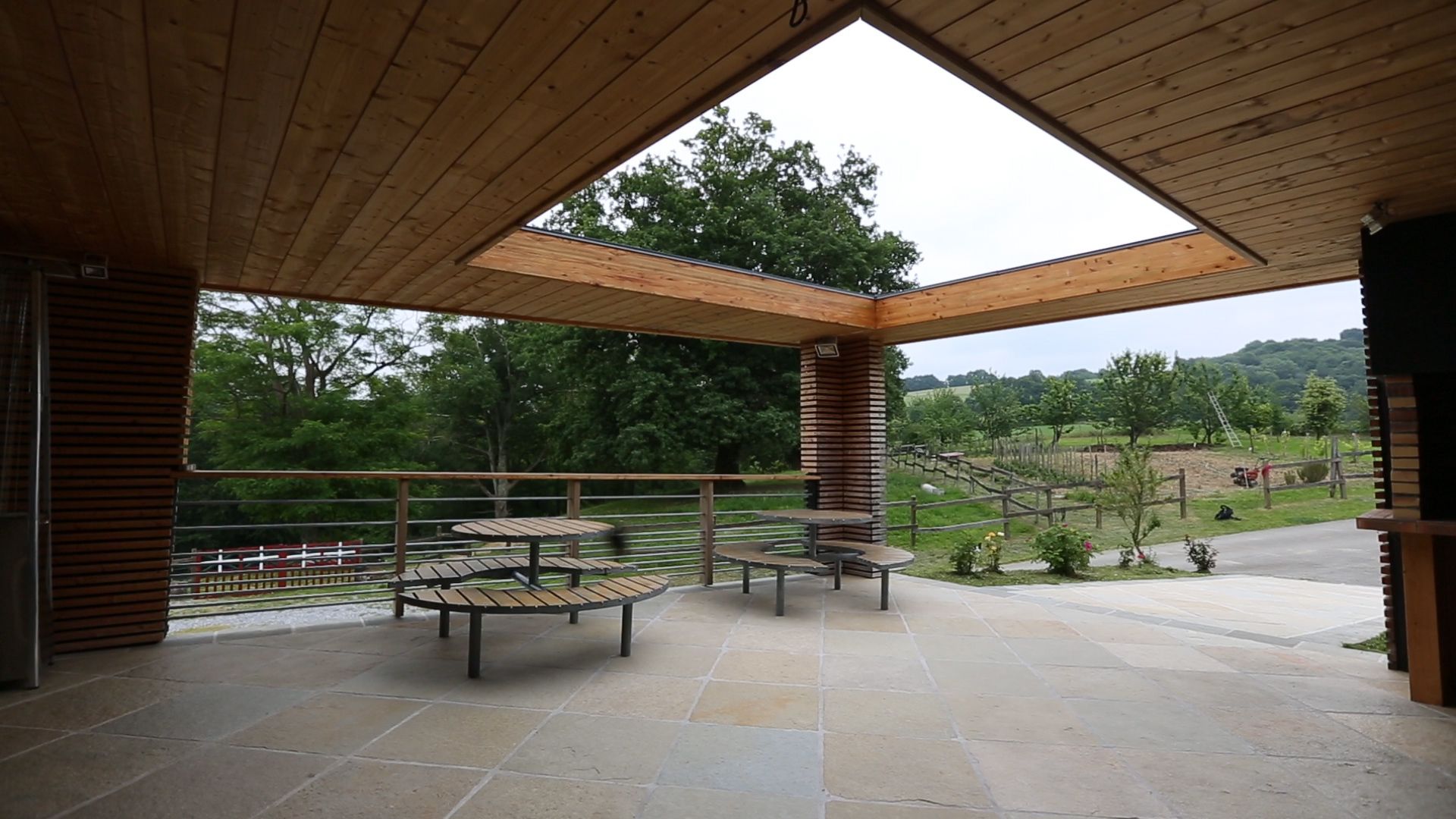 An outdoor patio with stone flooring, wooden tables and benches, and a view of a rural landscape with trees and a fence.