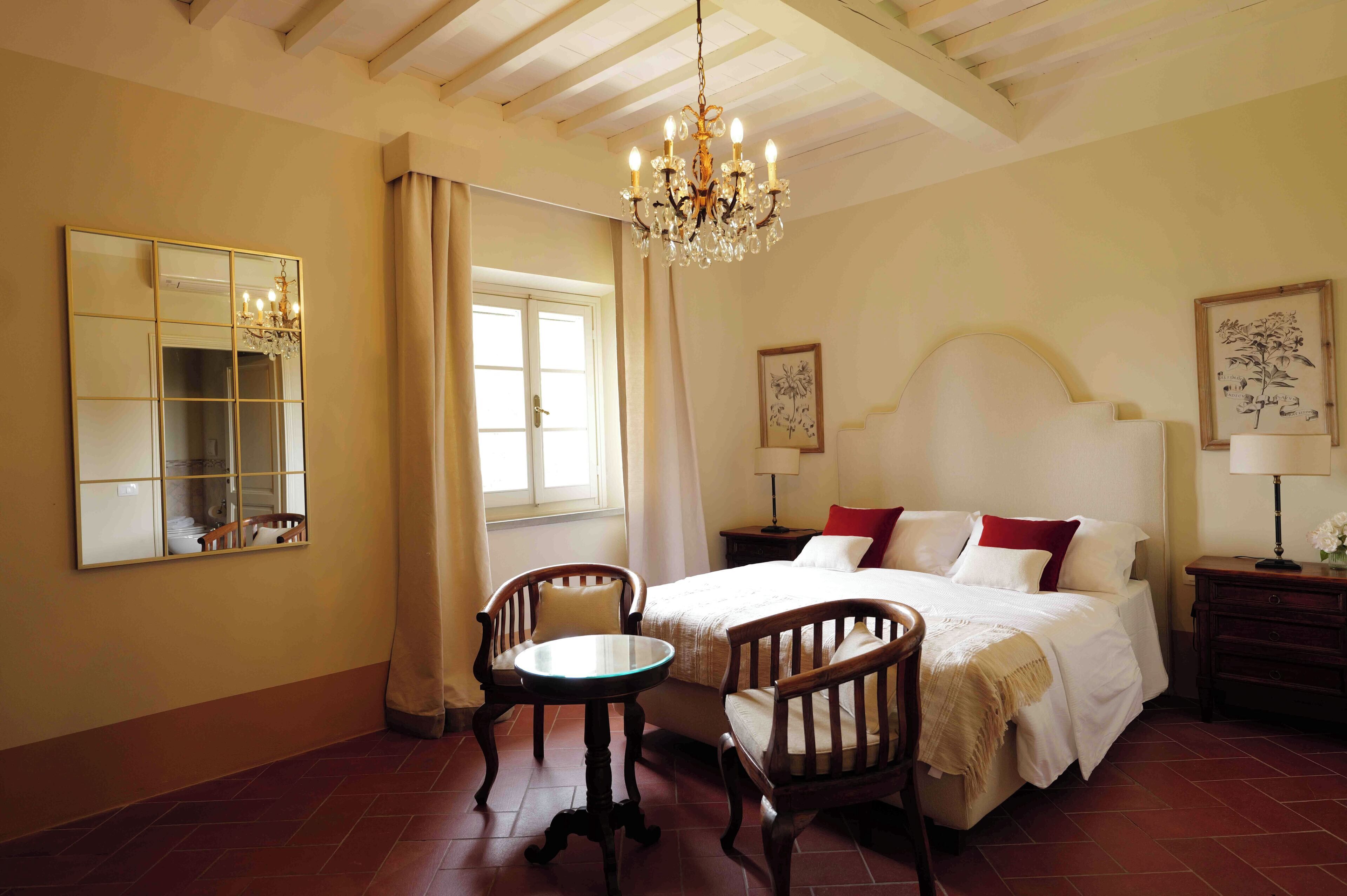 A bedroom with a bed, two chairs, a table, a chandelier, a mirror, and artwork on the walls.