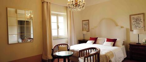 A bedroom with a bed, two chairs, a table, a chandelier, a mirror, and artwork on the walls.