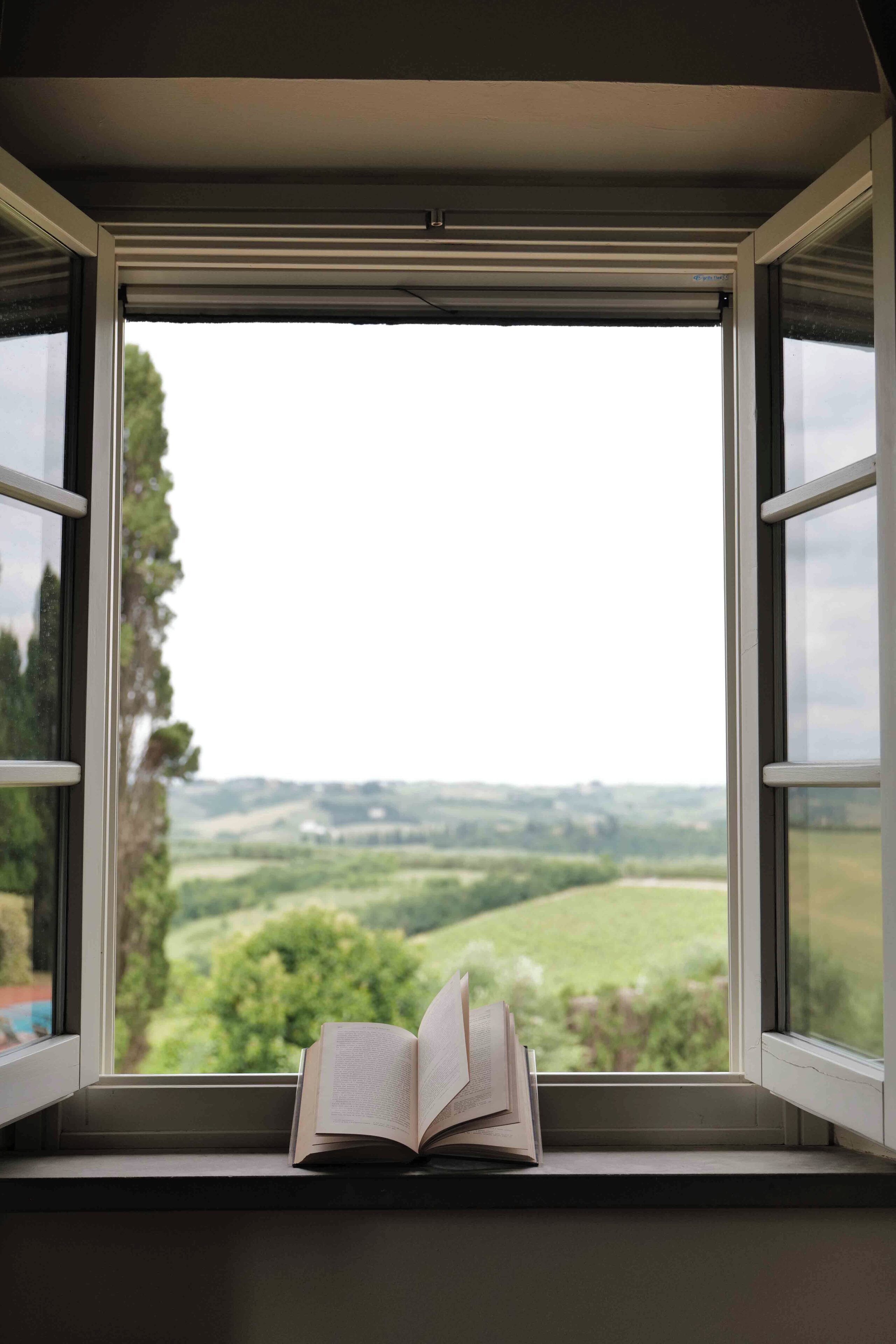 An open book on a windowsill with a view of a green landscape.