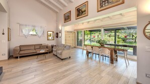 Amplia sala de estar con piso de madera, un sofá, una mesa de centro y un comedor con sillas y una mesa. Grandes ventanas ofrecen vistas a un jardín.