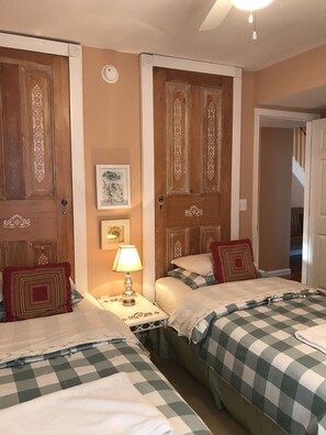 A bedroom with two beds, each with checkered bedding and decorative pillows. There is a nightstand with a lamp between the beds. The room has wooden doors with intricate designs and a framed picture on the wall.