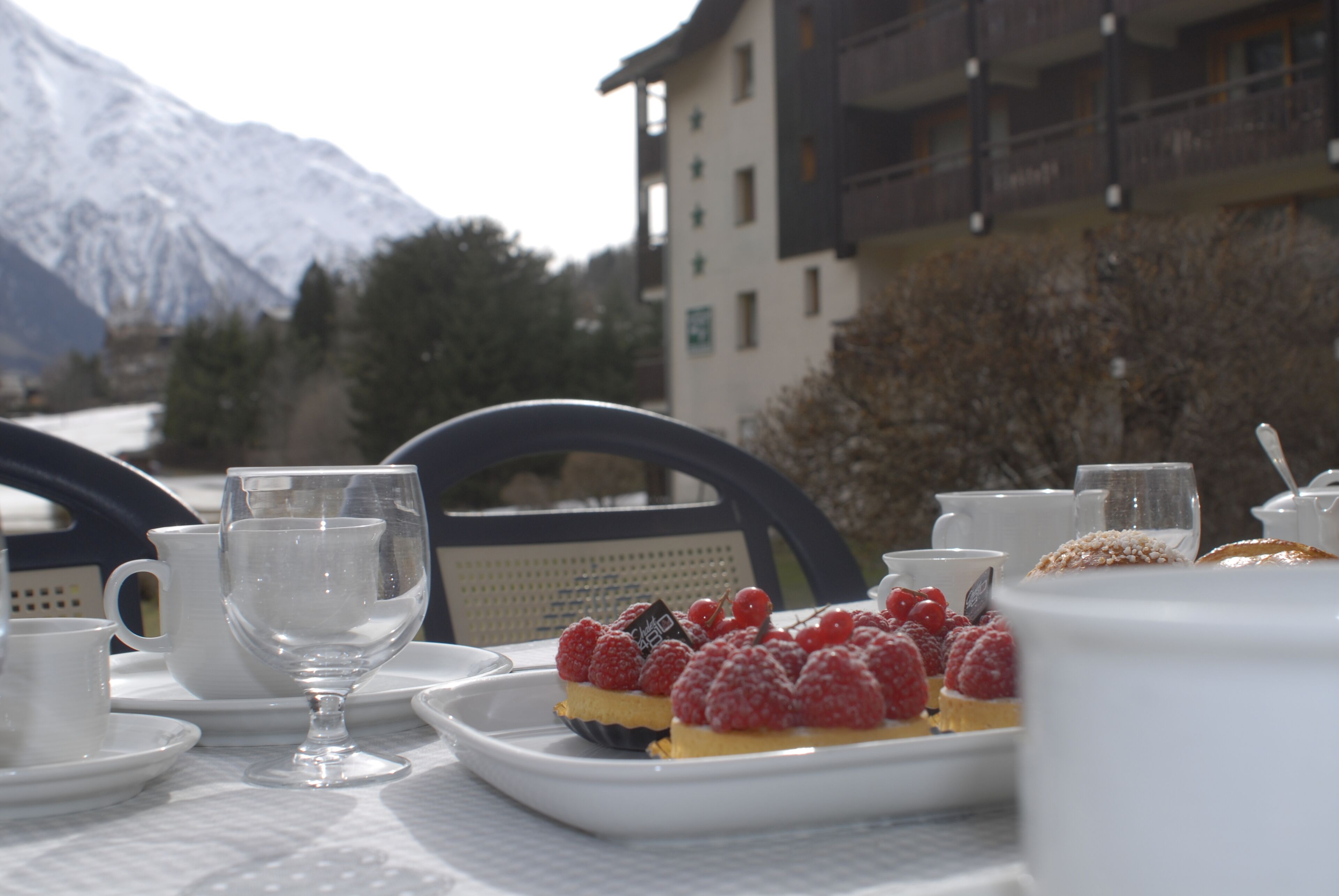 A table set with white dishes, a glass, and a dessert with raspberries, with a mountain in the background.