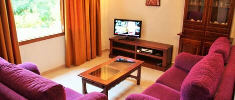 A living room with purple sofas, a glass coffee table, a TV on a wooden stand, and a cabinet.