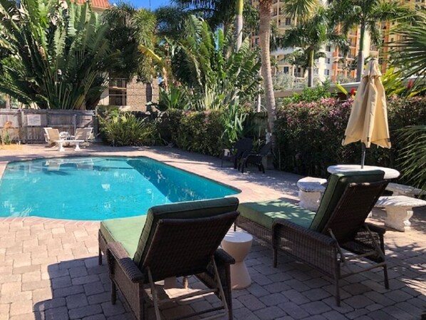 A swimming pool with lounge chairs and umbrellas, surrounded by lush greenery and a building in the background.
