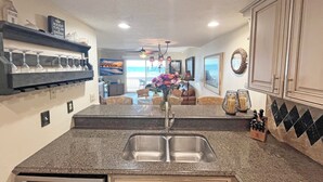 A kitchen with a granite countertop, stainless steel sink, and a dining area with a table and chairs. - Crescent Beach I-14 Four Winds Condo (St Augustine)