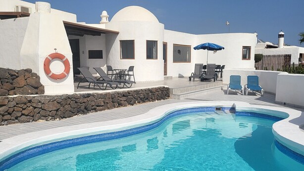 A clear blue swimming pool with white tiled edges, surrounded by white buildings with blue accents, and outdoor seating areas with umbrellas. - Amazing panoramic sea views!!! (Playa Blanca)
