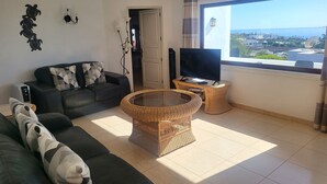 A living room with a sofa, a glass-top coffee table, a TV on a stand, and a view of the city through a large window. - Amazing panoramic sea views!!! (Playa Blanca)