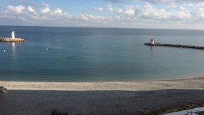 Vista costera con una playa de guijarros, un faro y un pequeño muelle que se adentra en el agua.