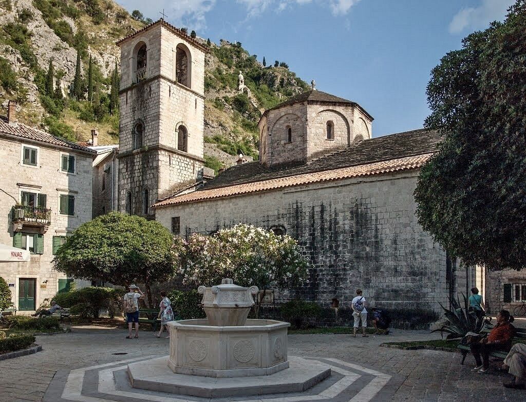 A courtyard with a fountain, stone buildings, and a bell tower.