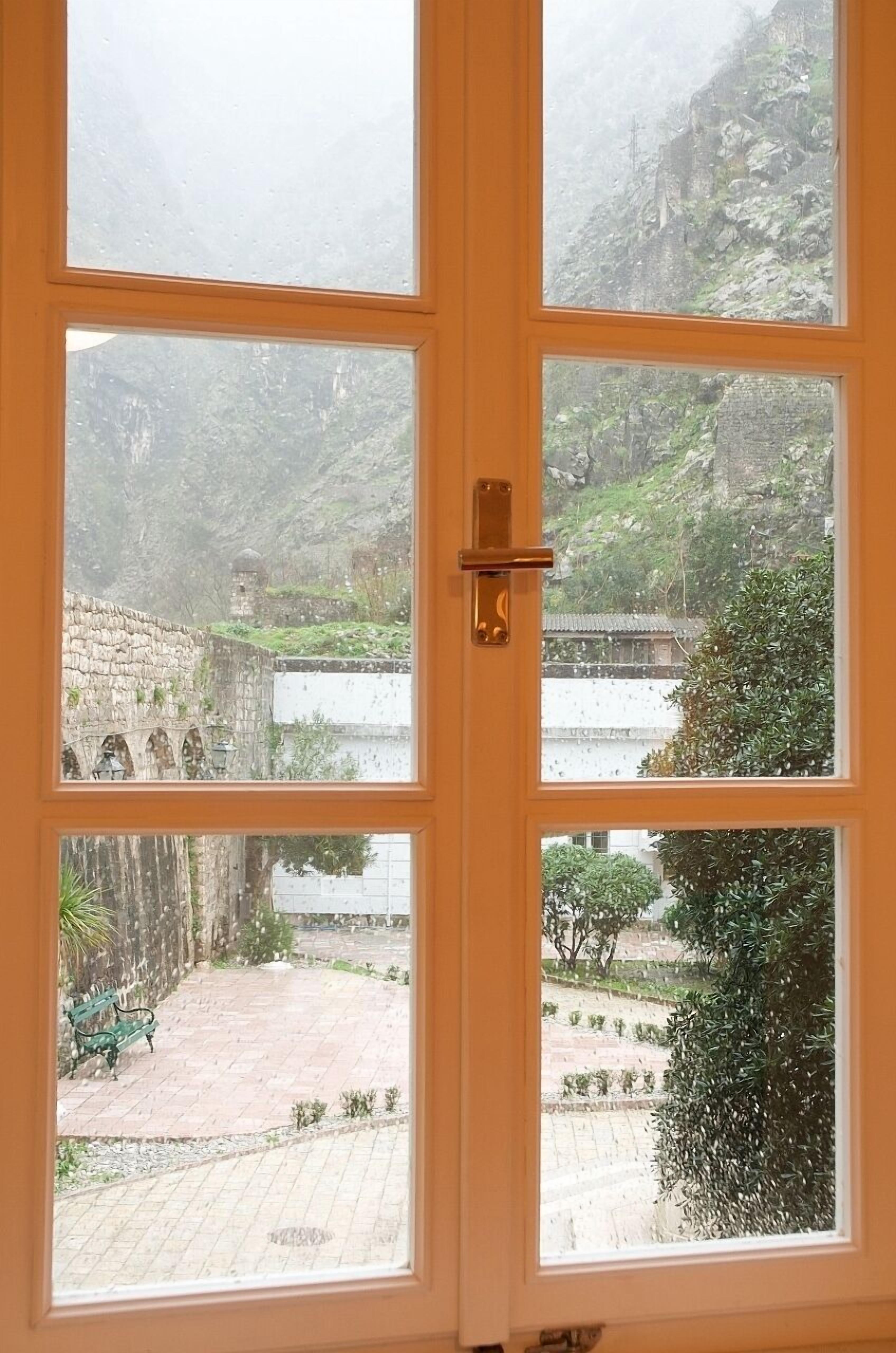 Une vue à travers une fenêtre à cadre en bois, donnant sur une cour intérieure avec un mur de pierre, un banc et un paysage montagneux en arrière-plan.