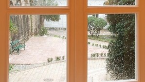 Une vue à travers une fenêtre à cadre en bois, donnant sur une cour intérieure avec un mur de pierre, un banc et un paysage montagneux en arrière-plan.