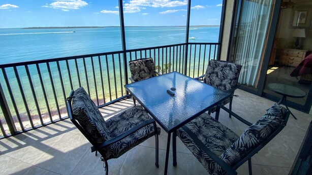 Un balcone con un tavolino in vetro e quattro sedie a motivi, con vista su un mare azzurro e una costa lontana.