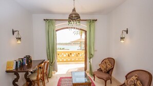 Ein Zimmer mit Esstisch, Stühlen, einer Couch und einem kleinen Tisch. Durch eine Tür ist ein Blick auf einen Außenbereich mit Zaun und Palmen.