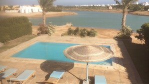 Ein Poolbereich mit Liegestühlen und Sonnenschirm, mit Blick auf ein Gewässer und eine entfernte Küstenlinie mit Gebäuden.