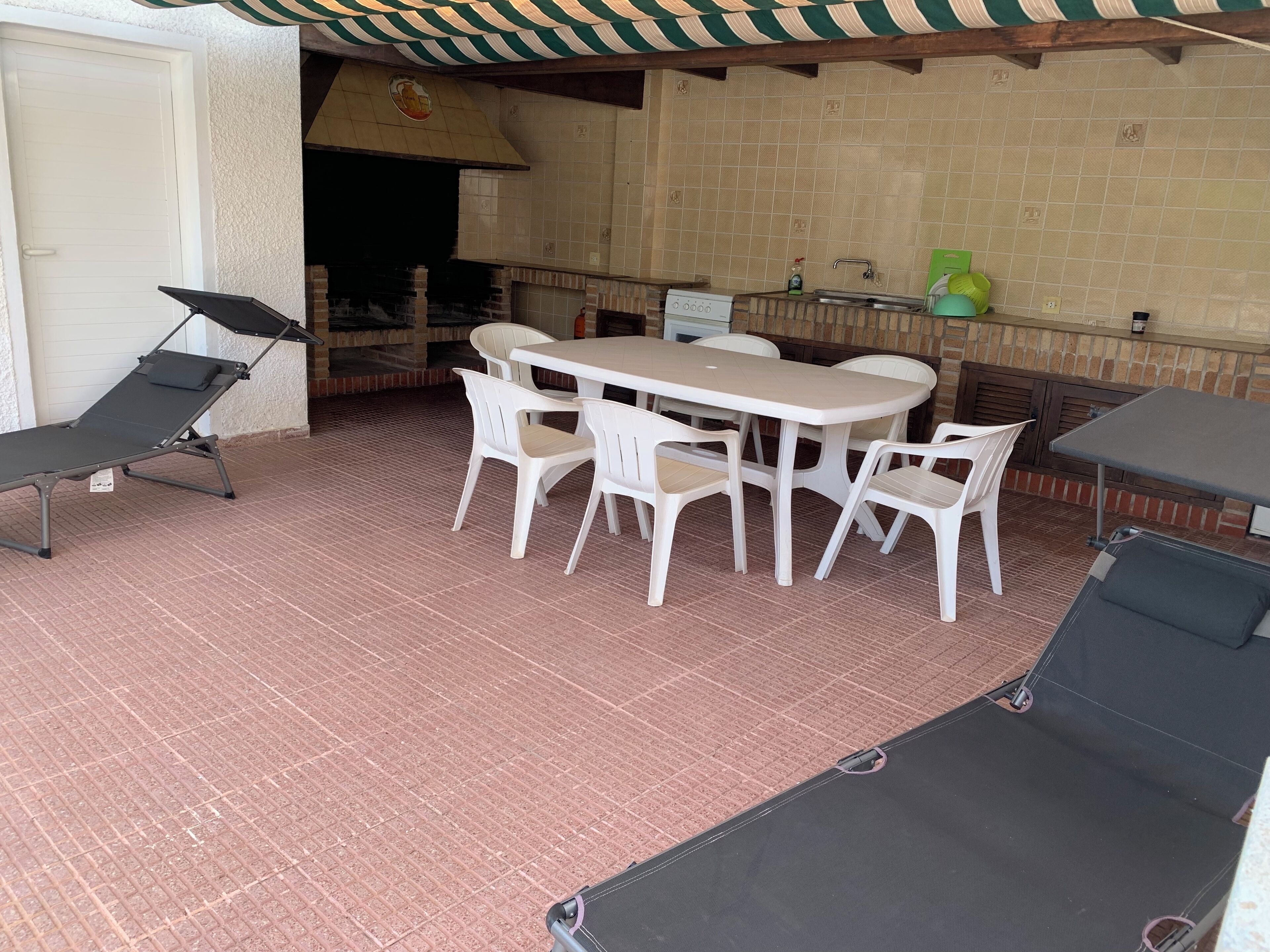 Un patio al aire libre con piso de ladrillo, una mesa blanca con sillas, una silla lounge negra y una zona de parrillada.