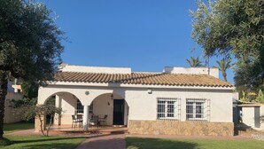 Un bâtiment doté d’un toit recouvert de tuiles, de murs blancs et d’un chemin pavé menant à un patio couvert avec des sièges extérieurs.