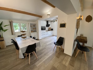 Un coin repas avec une table dressée pour quatre personnes, entourée de chaises. On aperçoit une cuisine en arrière-plan, avec des meubles blancs et un four noir.