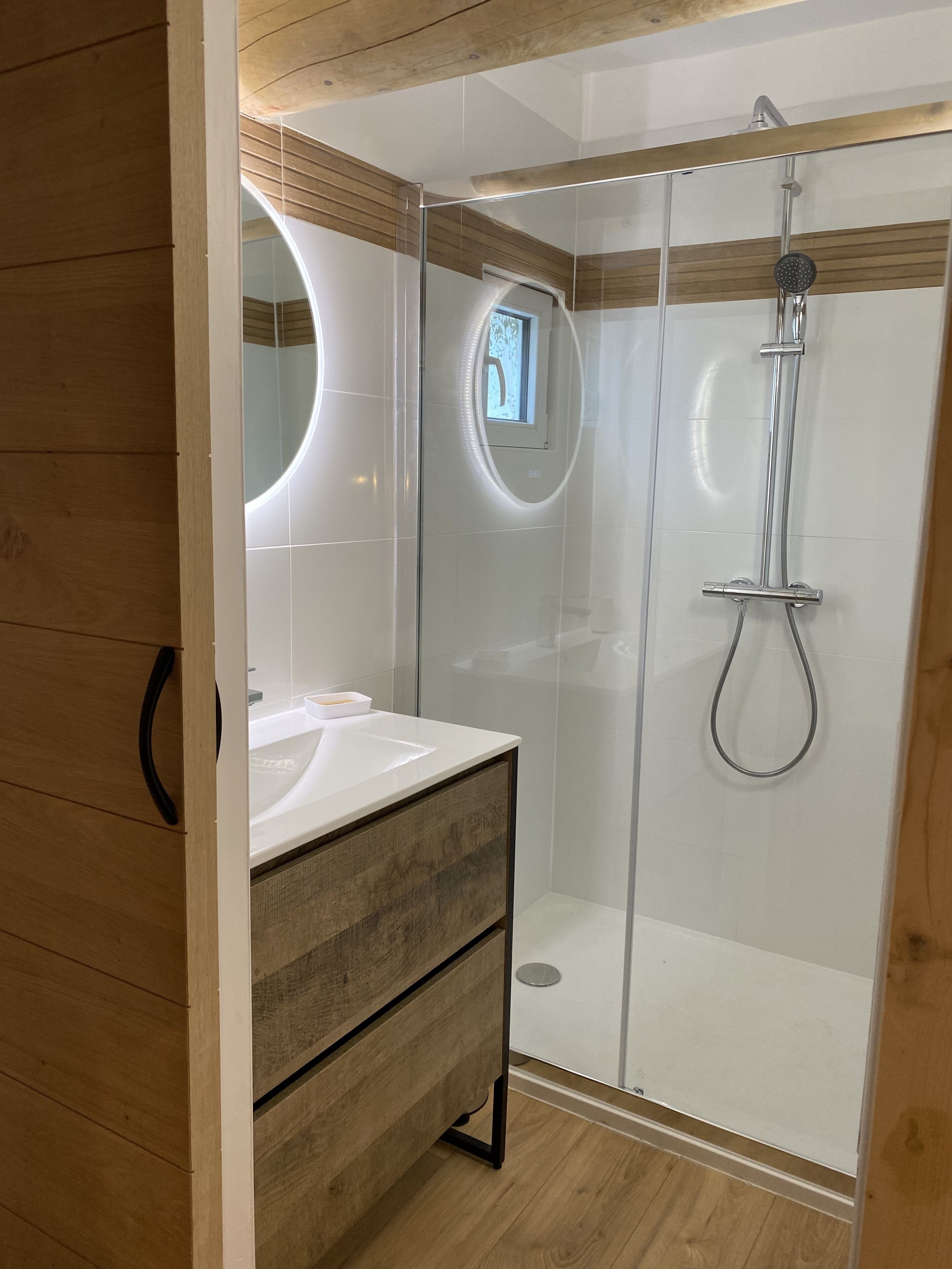 Een moderne badkamer met een douchecabine van glas, een houten wastafelmeubel en een spiegel met ronde verlichting.