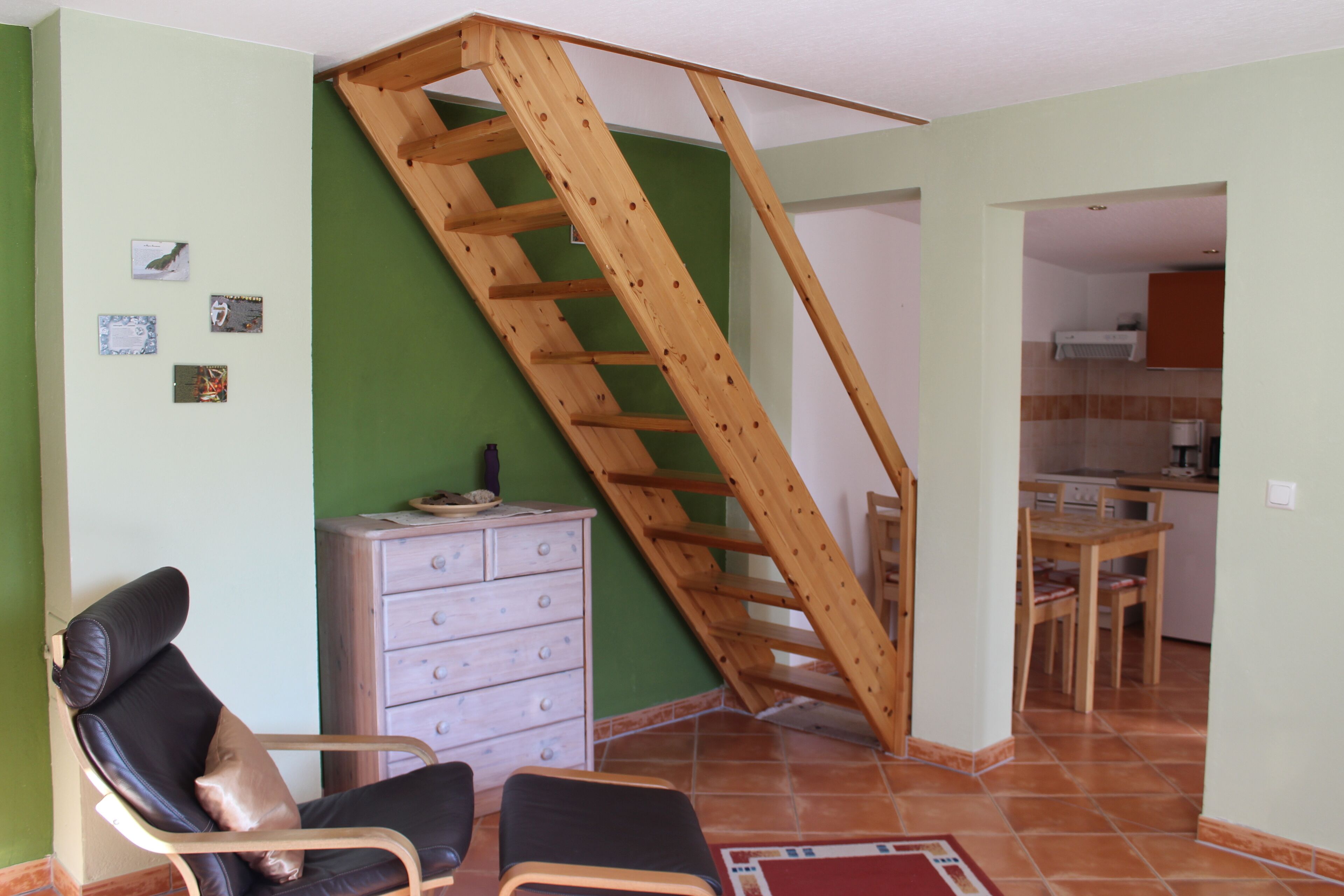 Una habitación con una escalera de madera, una silla, un aparador y una cocina visible a través de un vano abierto.