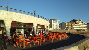 An outdoor café with orange chairs and tables, a concrete barrier, and buildings in the background. - fully equipped apartment on the first floor (capbreton)