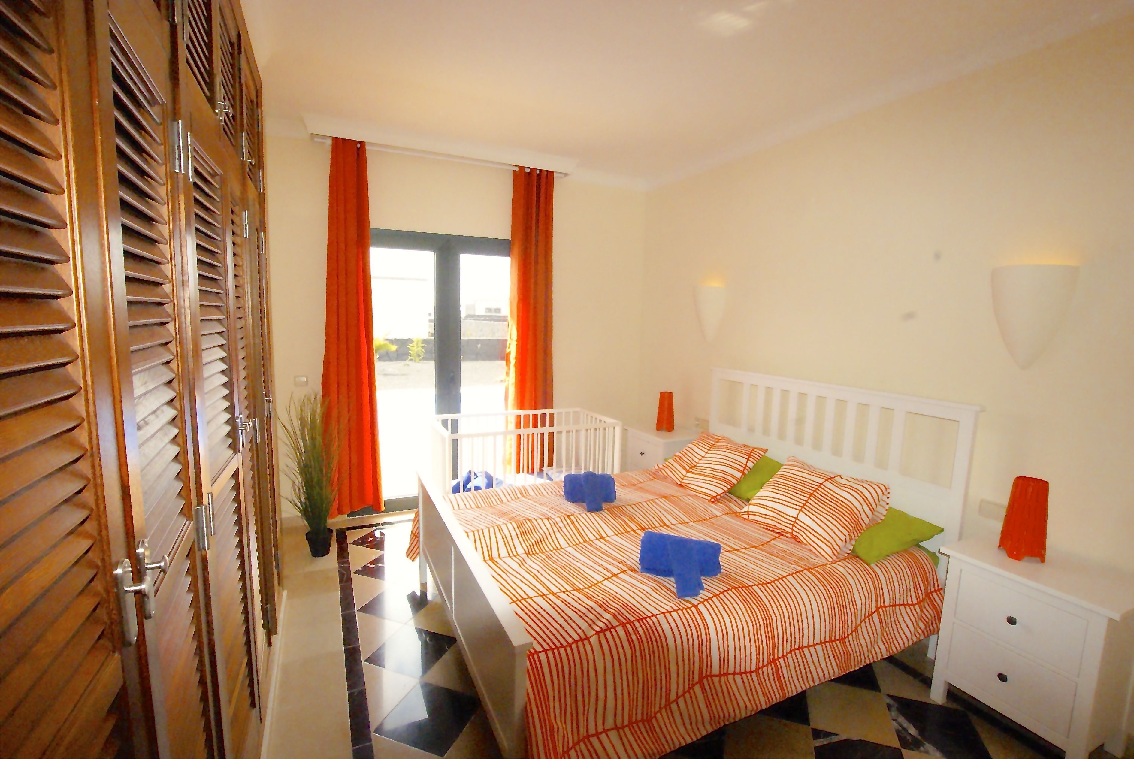 Camera da letto con un letto, un comodino bianco e vista attraverso una finestra con tende arancioni.