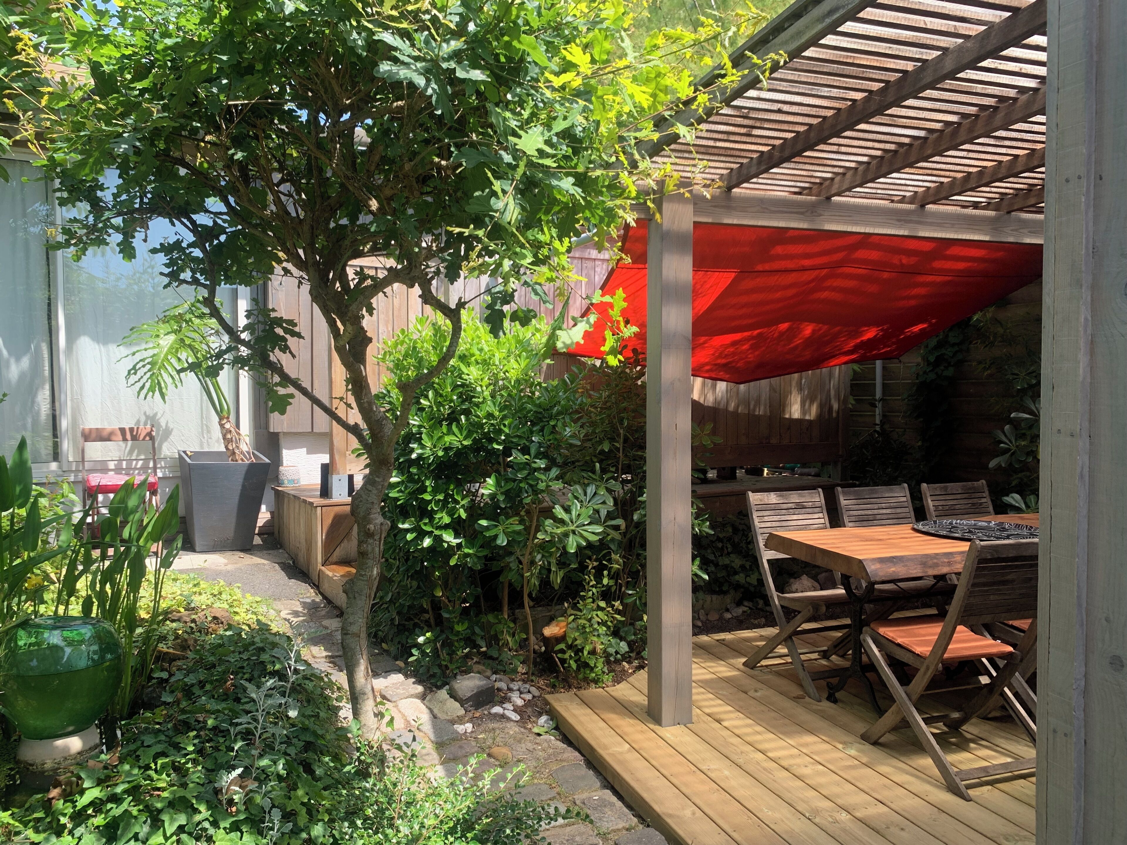 Een houten terras met een tafel en stoelen, omgeven door groen en een rood zonnescherm.