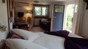 Quarto com cama, escrivaninha e cadeira. Há uma janela com cortinas, uma porta e vista para o verde lá fora.